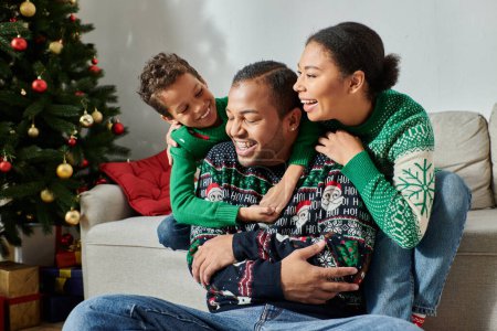 glückliche afrikanisch-amerikanische Familie in kuscheligen Pullovern, die sich warm umarmen und freudig lächeln, Weihnachten