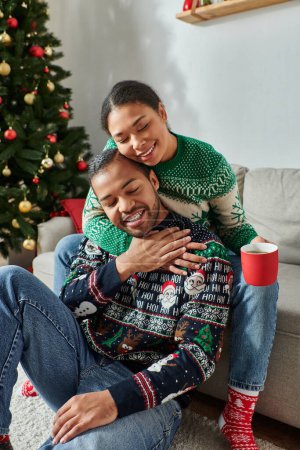 vertikale Aufnahme eines glücklichen afrikanisch-amerikanischen Paares in warmen Pullovern, das sich mit geschlossenen Augen umarmt, Weihnachten