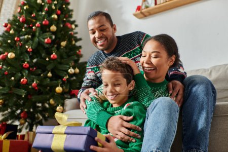 alegres padres afroamericanos abrazando a su hijo feliz sosteniendo un gran regalo de Navidad envuelto