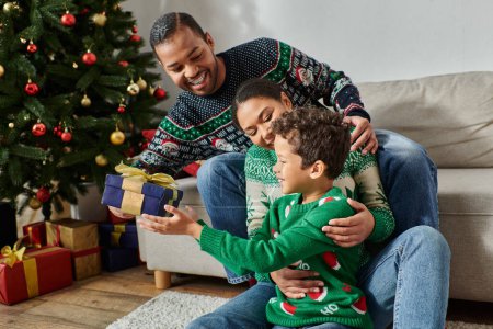 alegres padres cariñosos abrazando a su hijo sosteniendo presente y sonriendo alegremente, Navidad