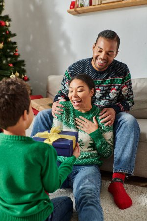 vertical shot of jolly african american parents hugging and receiving gift from their son, Christmas