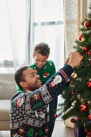 Senkrechte Aufnahme eines fröhlichen afrikanisch-amerikanischen Mannes, der zusammen mit seinem Sohn den Weihnachtsbaum schmückt