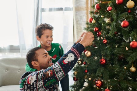 feliz afroamericano padre e hijo vistiendo suéteres acogedores decorando árbol de Navidad con chucherías