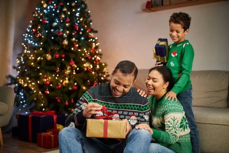 happy african american man unpacking present while his wife and son smiling at him, Christmas