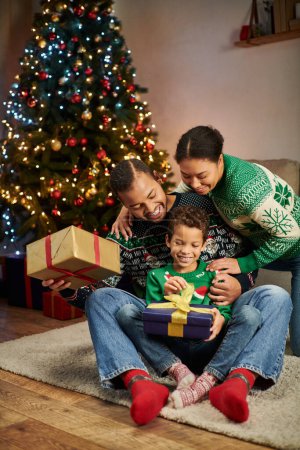 vertikale Aufnahme einer fröhlichen afrikanisch-amerikanischen Familie, die am Weihnachtsbaum sitzt und sich herzlich umarmt