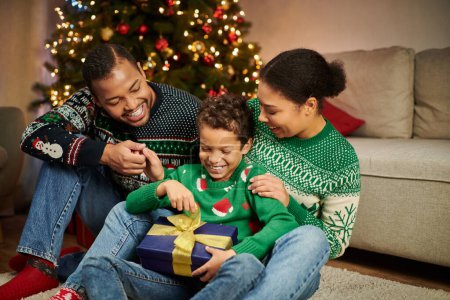 padres afroamericanos felices en suéteres acogedores mirando cómo su hijo desempacar regalo de Navidad