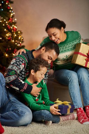 vertikale Aufnahme fröhlicher afrikanisch-amerikanischer Eltern, die ihren Sohn mit Weihnachtsgeschenk betrachten