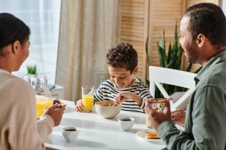 focus on happy african american boy enjoying breakfast and his blurred parents looking at him