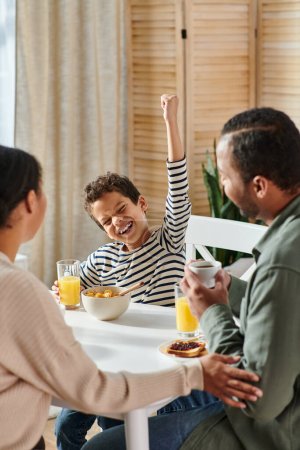 vertikale Aufnahme eines kleinen afrikanisch-amerikanischen Jungen, der beim Frühstück jubelt, während seine Eltern ihn anschauen