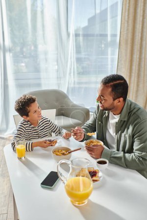 vertikale Aufnahme eines fröhlichen afrikanisch-amerikanischen Vaters und Sohnes, die das Frühstück genießen und glücklich lächeln
