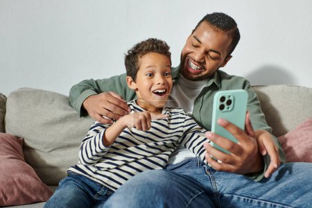 glücklich überrascht afrikanischer amerikanischer Junge schaut überrascht auf Handy, das auf dem Schoß seines Vaters sitzt