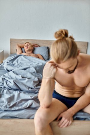 frustrierter bärtiger schwuler Mann sitzt mit gesenktem Kopf in der Nähe von Liebespartner, der im Schlafzimmer schläft, Konflikt