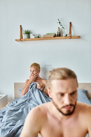 worried gay man covering mouth with hand near offended boyfriend on blurred foreground in bedroom