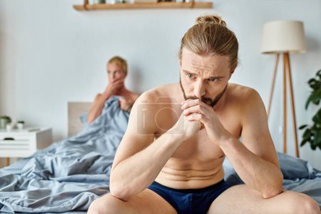 Enttäuschter schwuler Mann in Unterhosen sitzt neben Freund auf verschwommenem Hintergrund im Schlafzimmer