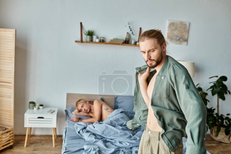 Frustrierter bärtiger schwuler Mann verkleidet sich in der Nähe von Freund, der morgens im Schlafzimmer schläft, Liebeskonflikt