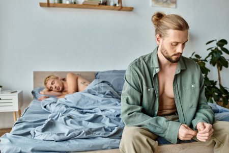 Téléchargez les photos : Offensé barbu gay l'homme assis et habiller près copain couché dans chambre le matin - en image libre de droit