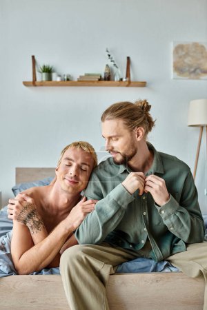 happy gay man leaning on smiling bearded boyfriend sitting in modern bedroom, happiness