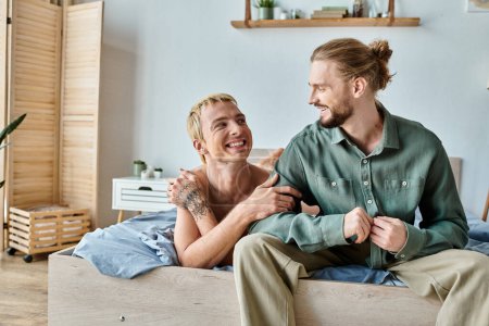 joyful tattooed gay man looking at smiling bearded boyfriend sitting in bedroom, happy relationship