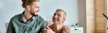 joyful tattooed gay man looking at smiling bearded boyfriend sitting in bedroom, horizontal banner