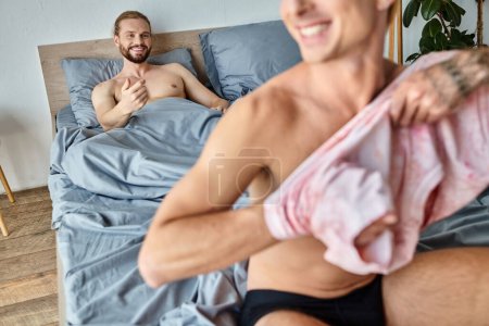 joyful bearded gay man lying and smiling near boyfriend dressing up in bedroom, happy morning scene