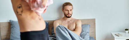 Téléchargez les photos : Bouleversé barbu gay l'homme assis sur lit près de l'amour partenaire habillage jusqu 'dans chambre à coucher, horizontal bannière - en image libre de droit
