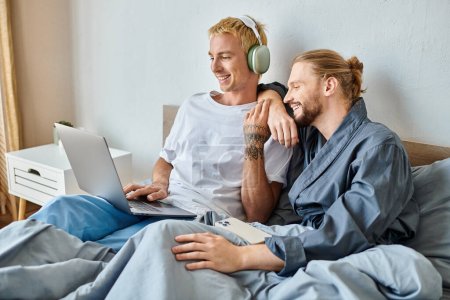 alegre gay hombre en auriculares usando portátil cerca barbudo novio con smartphone en dormitorio