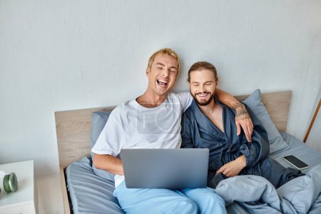 fröhliches homosexuelles Paar schaut Film auf Laptop und lacht im Bett, glückliche Liebe und Beziehungen