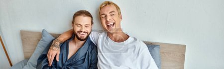 cheerful gay couple laughing while sitting on bed in morning, happy relationships, horizontal banner