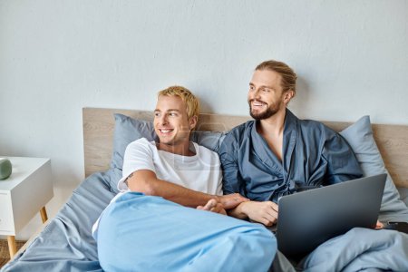 happy gay couple looking away while watching movie on laptop in bedroom, leisure and tranquility