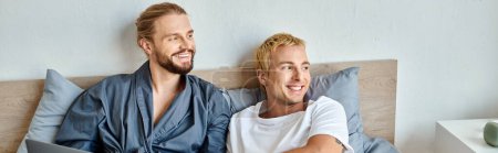 happy gay couple smiling and looking away while sitting in bedroom in morning, horizontal banner