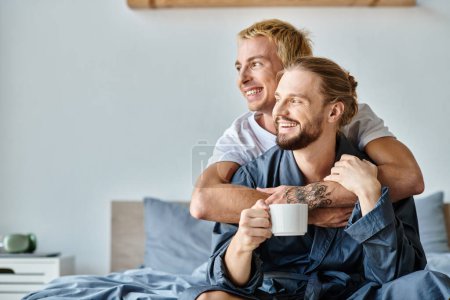 joyful tattooed gay man embracing bearded boyfriend with coffee cup in bedroom, romantic scene