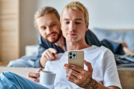 tätowierter schwuler Mann mit Kaffeetasse surft im Internet auf dem Handy in der Nähe eines lächelnden Freundes im Schlafzimmer
