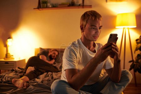 untreuer schwuler Mann chattet auf Handy in der Nähe von Liebespartner, der nachts im Schlafzimmer schläft