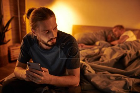 Photo for Disloyal bearded gay man browsing date app on smartphone near partner sleeping at night in bedroom - Royalty Free Image