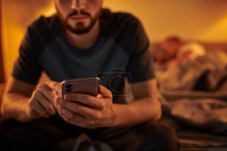 Photo for Cropped view of unfaithful gay man messaging on smartphone near partner sleeping at night in bedroom - Royalty Free Image
