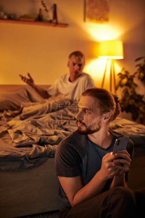 Unzufriedener schwuler Mann streitet mit Freund Nachrichten auf Smartphone in der Nacht im Schlafzimmer, Ärger