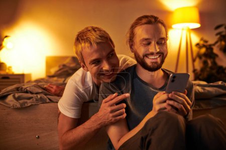 fröhlicher bärtiger schwuler Mann surft nachts im Internet auf dem Handy neben lächelndem Freund im Schlafzimmer
