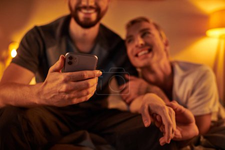 Lächelnder bärtiger Schwuler surft nachts im Internet auf dem Handy in der Nähe eines glücklichen Freundes im Schlafzimmer