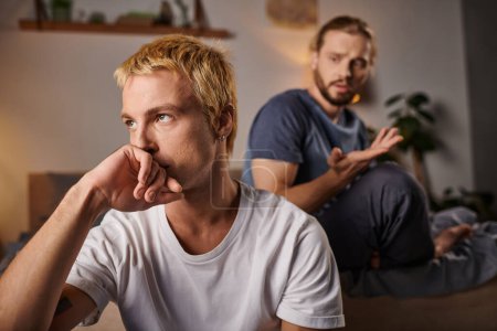 offended gay man holding hand near face and looking away near boyfriend at night in bedroom