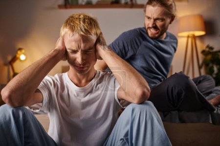 Photo for Stressed gay man covering ears with hands near angry boyfriend quarreling in bedroom at night - Royalty Free Image