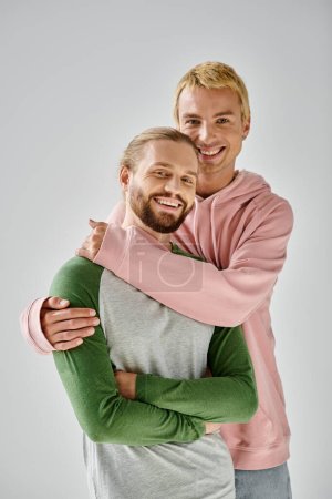 happy gay couple in stylish casual attire embracing and looking at camera on grey backdrop
