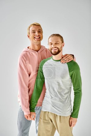 joyful gay couple in stylish casual attire standing and looking at camera on grey backdrop
