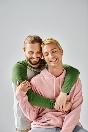 fröhlicher bärtiger schwuler Mann umarmt stilvollen Freund lächelnd mit geschlossenen Augen vor grauem Hintergrund