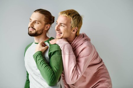 happy and dreamy gay couple in trendy casual attire looking away on grey backdrop, harmony and love