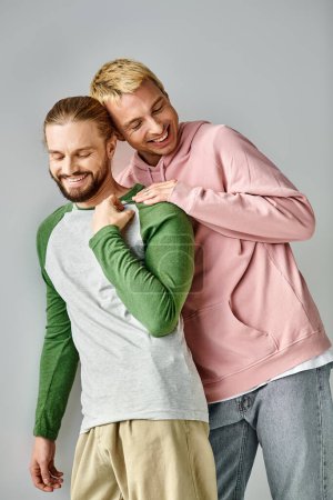 joyful gay couple in trendy casual attire posing with closed eyes on grey backdrop, love and harmony