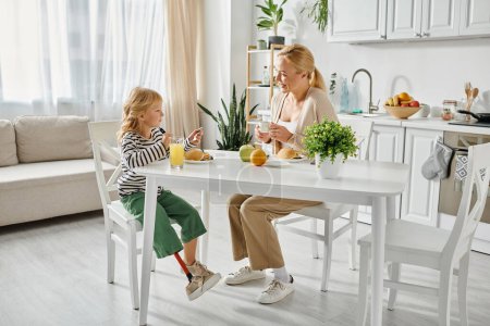 Téléchargez les photos : Fille préscolaire avec prothèse jambe petit déjeuner avec mère blonde heureuse dans la cuisine moderne - en image libre de droit