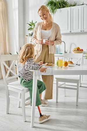 Foto de Mujer feliz de pie cerca de la hija con la pierna protésica desayunando en la cocina, inclusión - Imagen libre de derechos
