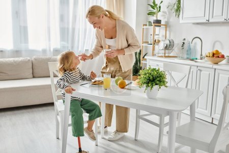Foto de Madre feliz de pie cerca de la hija con prótesis de pierna desayunando en la cocina, discapacidad - Imagen libre de derechos