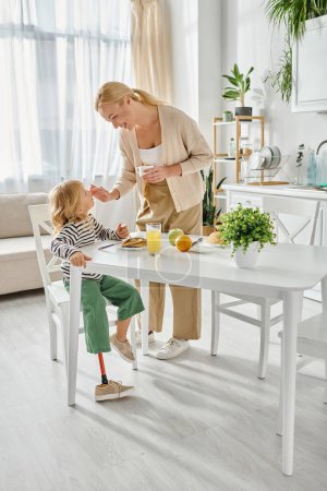 Téléchargez les photos : Mère heureuse debout près de la fille avec la jambe prothétique petit déjeuner dans la cuisine, adaptative - en image libre de droit