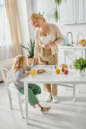 Téléchargez les photos : Mère heureuse debout près de fille mignonne avec jambe prothétique petit déjeuner dans la cuisine, inclusion - en image libre de droit